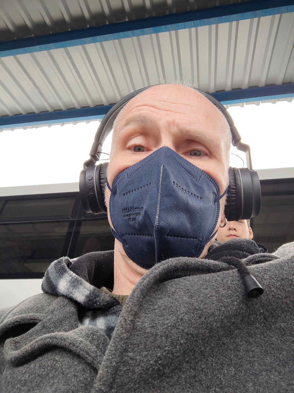 portrait of man wearing grey coat, black headphone, and blue face mask, male in the background, watching (but not prying; caption meant to be a joke)